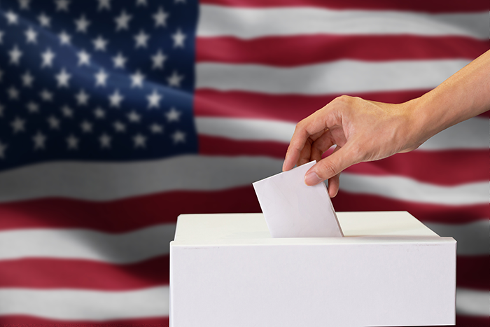 an image of a hand placing a piece of paper into a white box with an American flag in the background