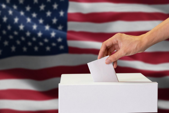 A person putting a ballot in a box in front of an American flag